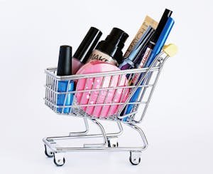 Miniature shopping cart filled with a variety of cosmetics on a white background.